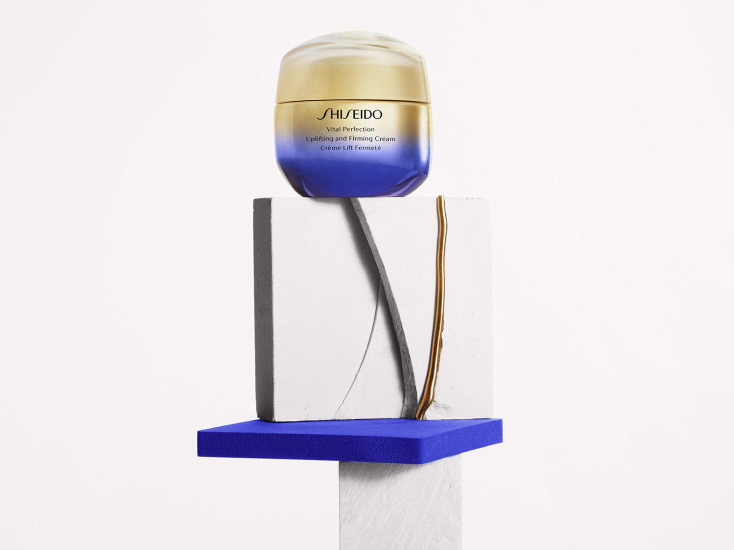Shiseido Vital Perfection Lifting and Firming Cream in einem Tiegel mit goldenem Deckel, der auf einem weißen Sockel mit blauen Akzenten steht.