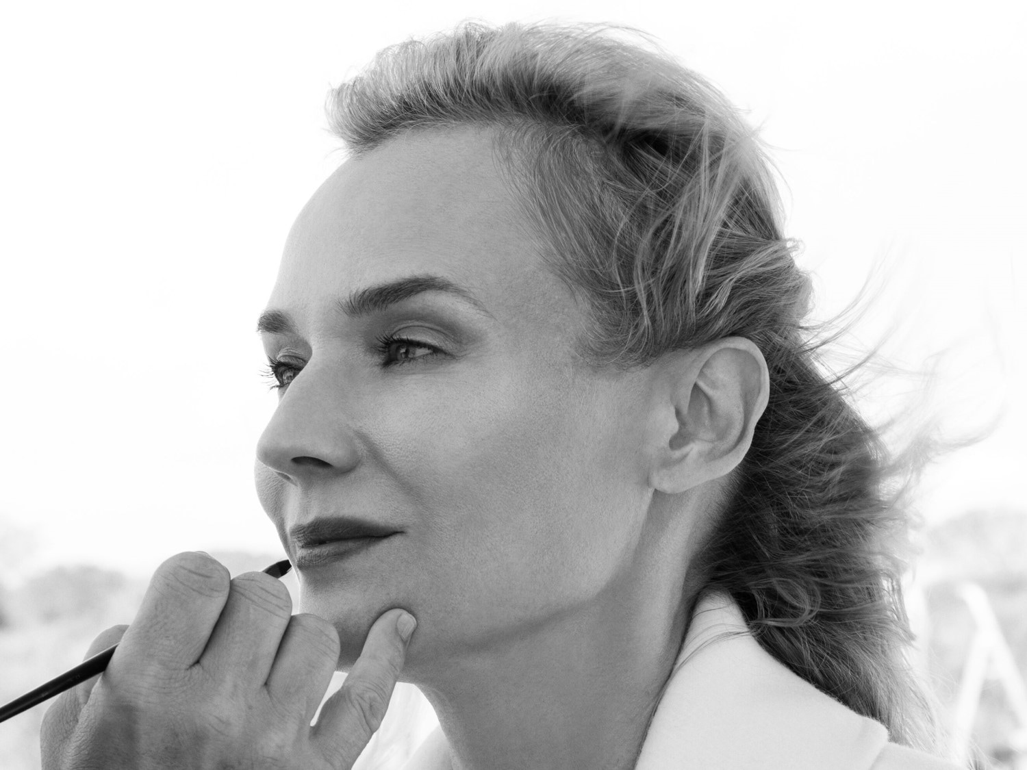 En primer plano, una mujer de perfil recibe un toque final de maquillaje con un pincel fino, resaltando sus labios con un color sutil mientras su mano descansa suavemente sobre su barbilla.