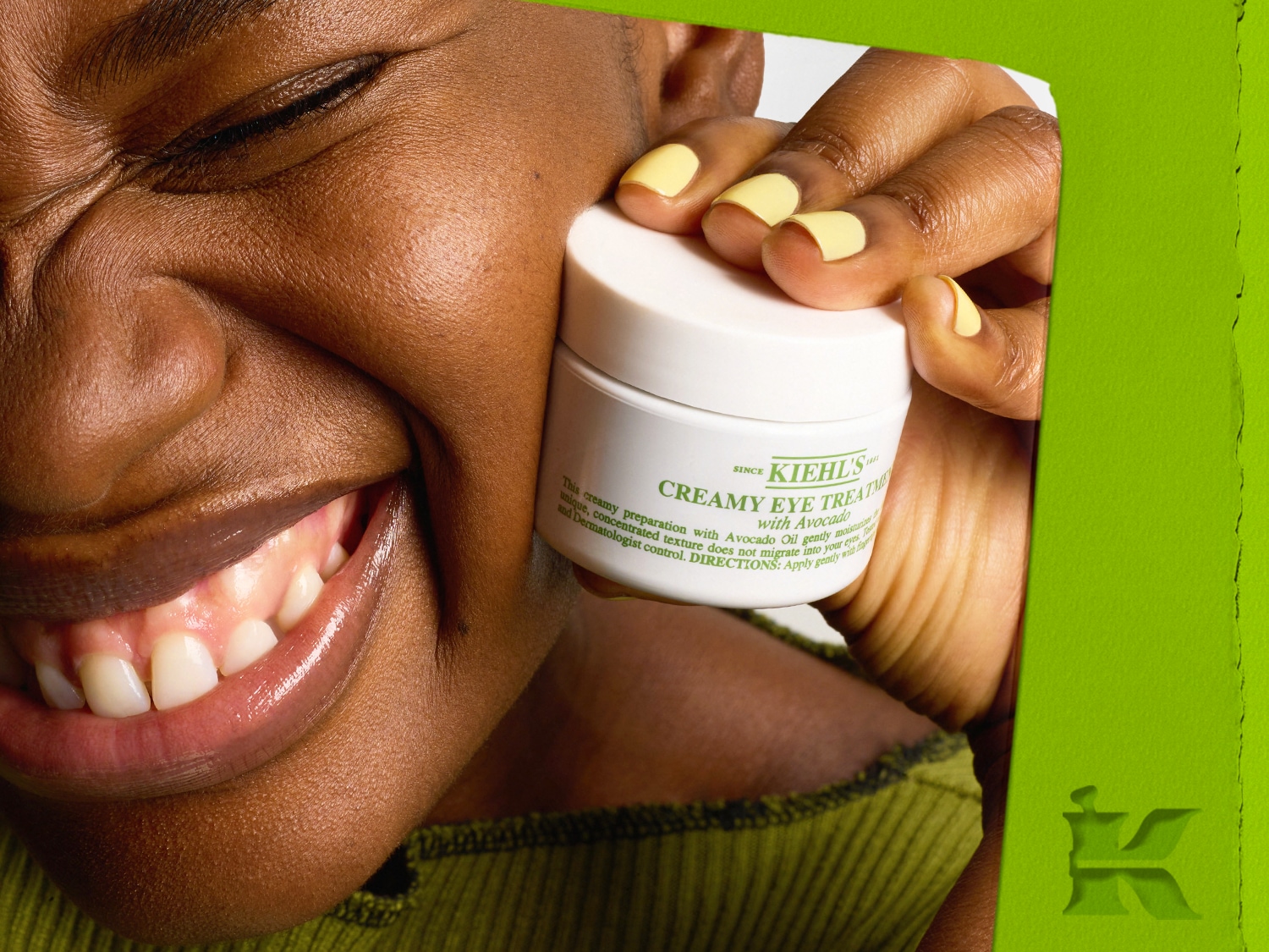 Egy mosolygós, sötét bőrű nő sárga körmökkel Kiehl's Creamy Eye Treatment krémet tartja, mely avokádóval készült, a hidratálásért és a szemkörnyék ápolásáért.
