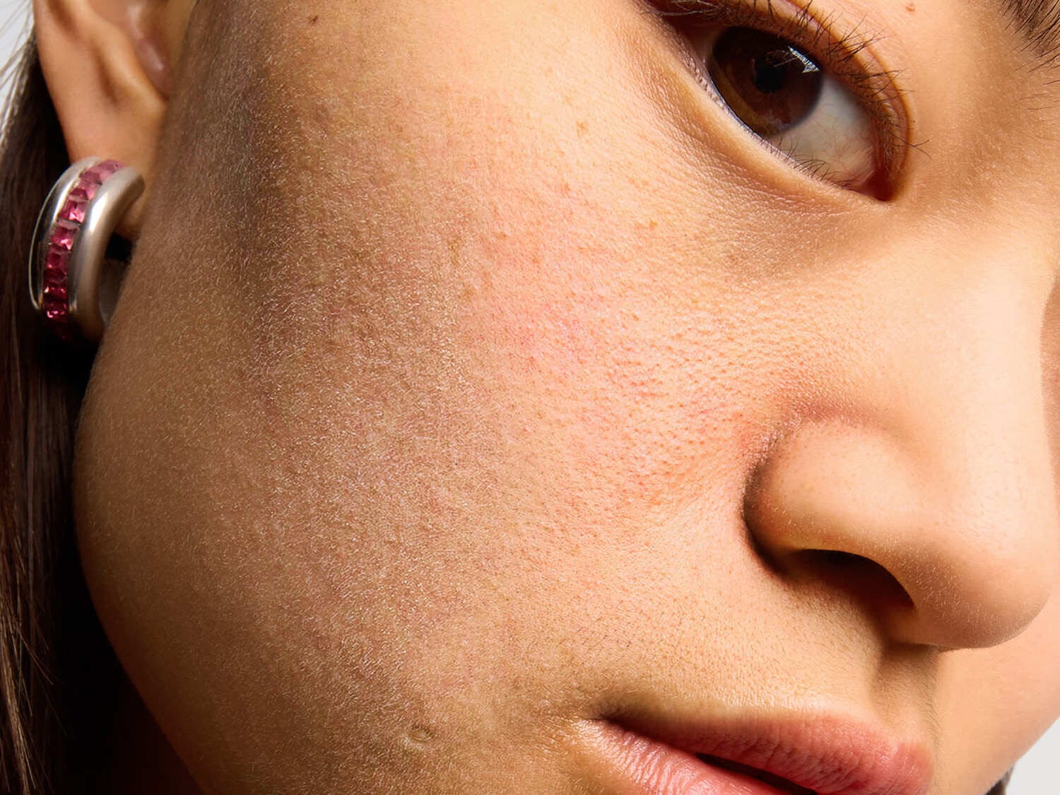 Nagyított felvétel egy nő arcáról, a fókusz a bőr textúráján, a szemén és egy ezüstszínű, rózsaszín kövekkel díszített fülbevalón van.