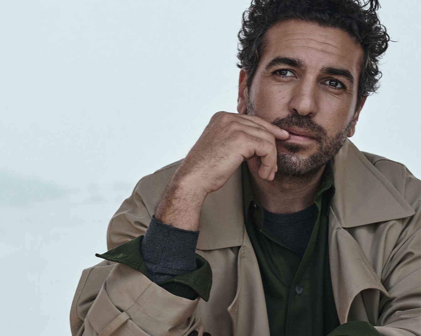 Ein Mann mit lockigem, dunklem Haar und Bart trägt einen beigen Trenchcoat über einem grünen Hemd und einem grauen Pullover, während er nachdenklich mit der Hand am Kinn posiert.
