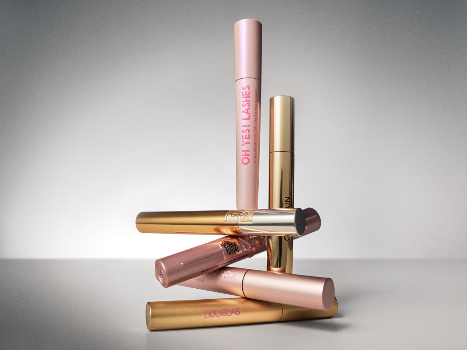 Een stapel mascara's in gouden en roze tinten, waaronder 'OH YES! LASHES' voor extra volume en 36 uur langhoudende mascara, en Douglas mascara.