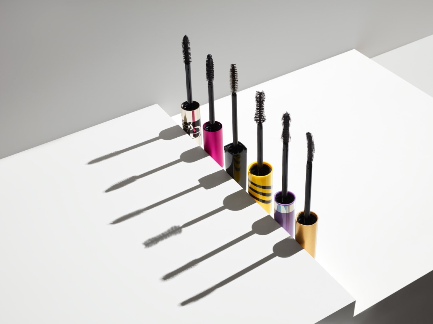 Zes kleurrijke mascara's staan op een witte achtergrond, elk met een zwarte borstel die uit de huls steekt, klaar om je wimpers te transformeren.