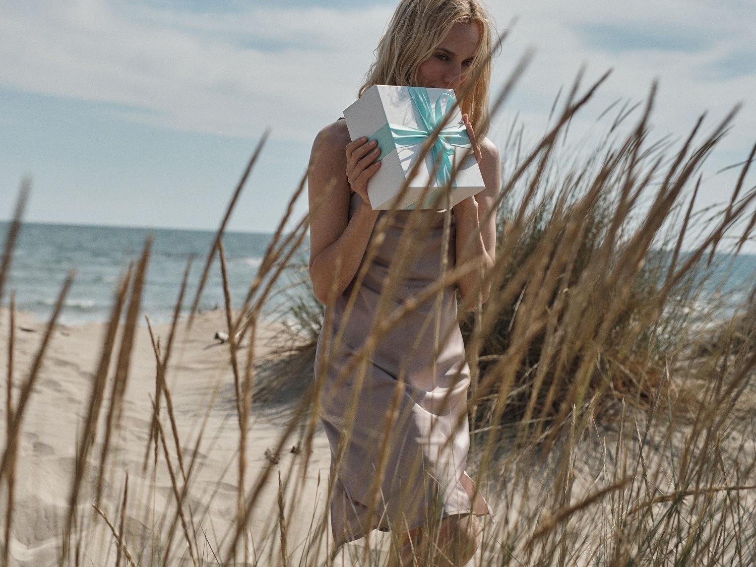 Sur une plage ensoleillée, une femme blonde tient un cadeau blanc orné d'un ruban bleu clair, évoquant la joie d'offrir et de recevoir.