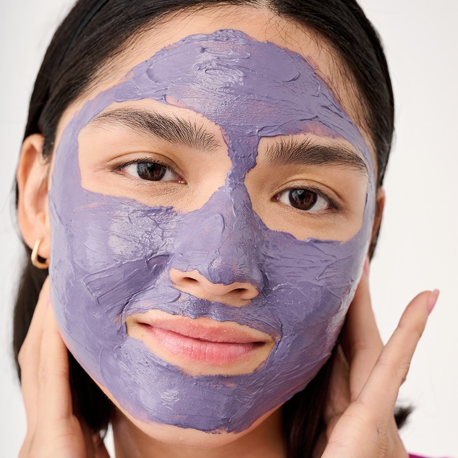 Nahaufnahme einer Frau mit einer lila Gesichtsmaske, die ihre Haut berührt und ein Gefühl von Entspannung und Hautpflege vermittelt.