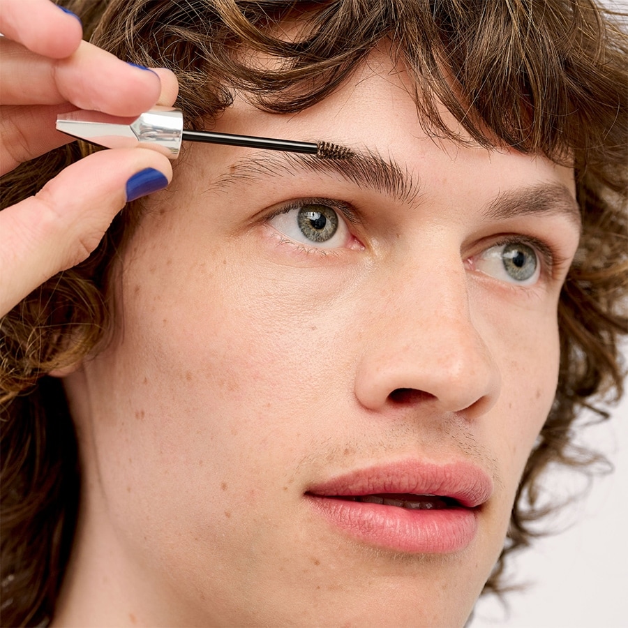 Ein Mann mit Sommersprossen und lockigem Haar erhält eine Augenbrauenbehandlung mit einer silbernen Bürste, um einen gepflegten Look zu erzielen.
