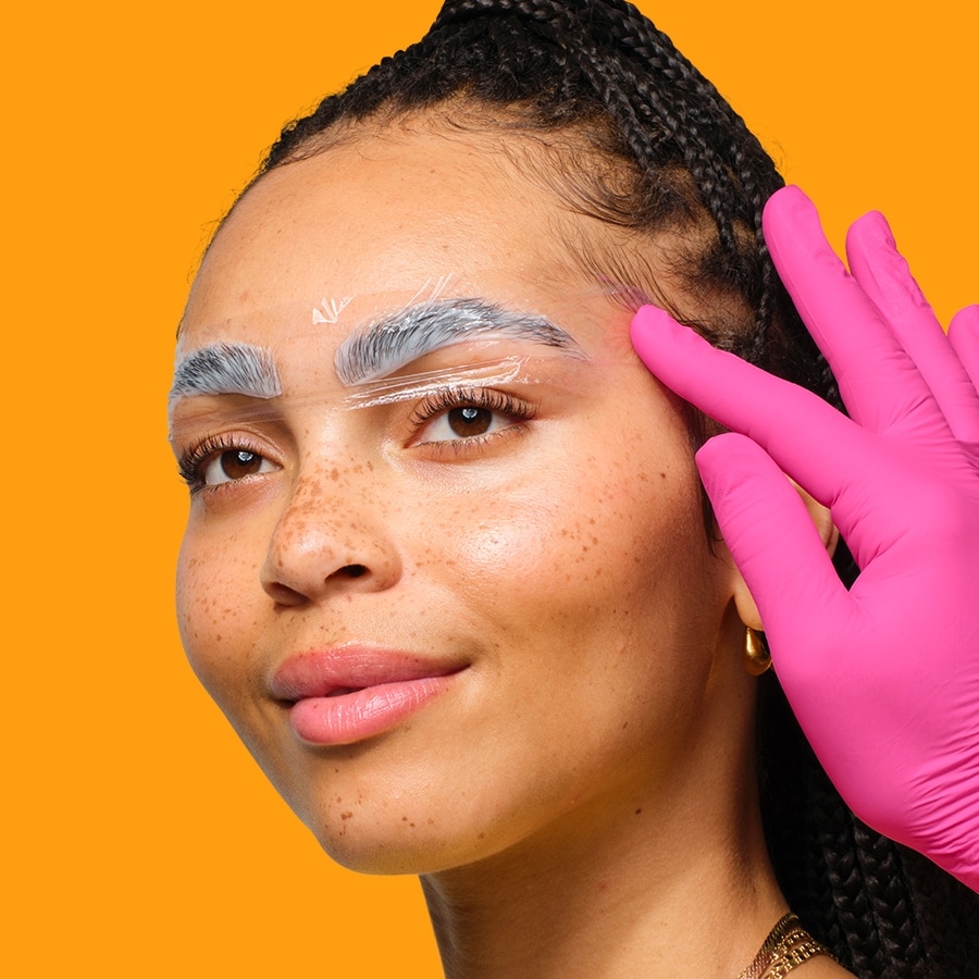 Nahaufnahme eines Frauengesichts mit gebleichten Augenbrauen, die mit einer transparenten Folie abgedeckt sind, während eine Hand in einem pinkfarbenen Handschuh die Folie andrückt.