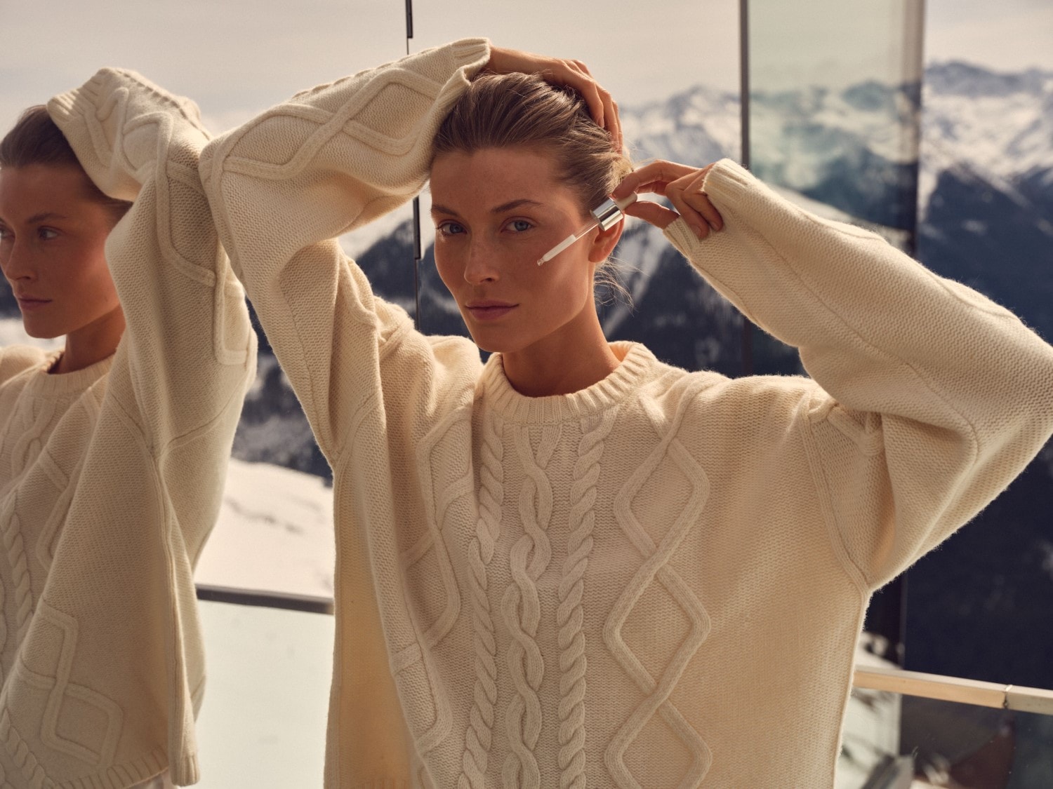 Een vrouw met een crèmekleurige trui brengt serum aan op haar gezicht met een pipet, terwijl ze in de spiegel kijkt met een berglandschap op de achtergrond.