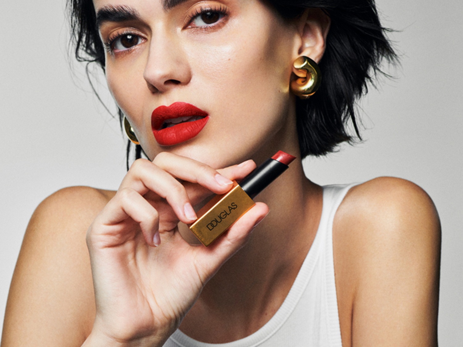 Kobieta z krótkimi czarnymi włosami i złotymi kolczykami trzyma w dłoni szminkę Douglas w złotym opakowaniu, prezentując intensywny czerwony odcień na ustach.