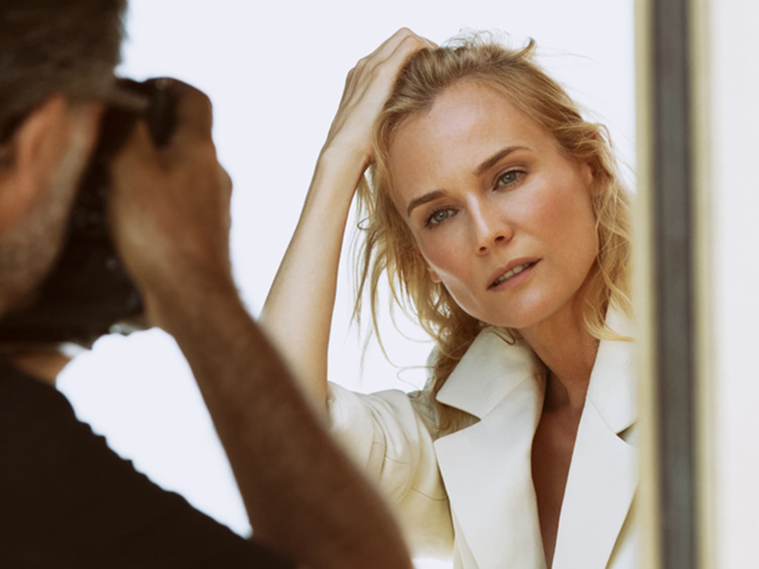Zdjęcie przedstawia blondwłosą kobietę w białej marynarce, pozującą fotografowi, z ręką we włosach i delikatnym makijażem.