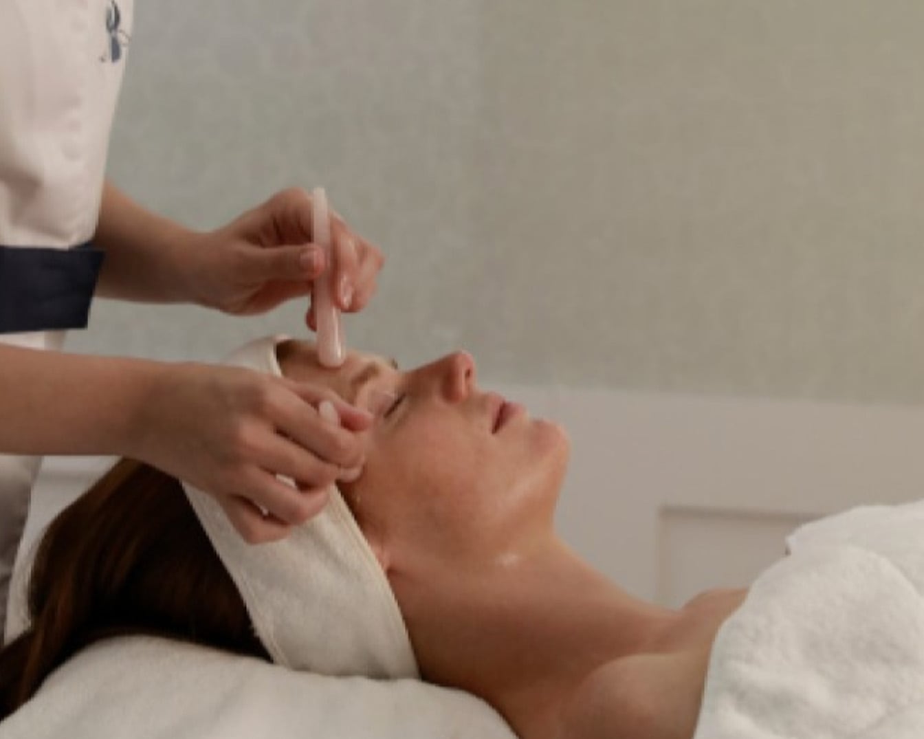 Kobieta relaksuje się podczas zabiegu na twarz z użyciem różowego wałka do masażu, leżąc z zamkniętymi oczami i opaską na włosach.