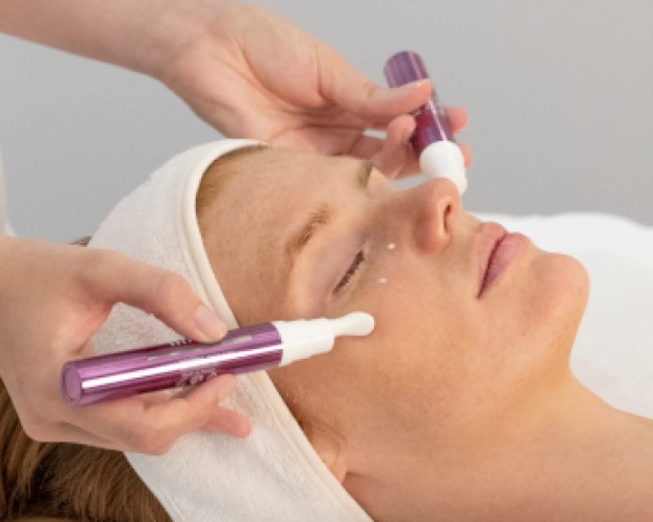 Kobieta z opaską na włosach otrzymuje zabieg kosmetyczny za pomocą dwóch fioletowych aplikatorów z białymi końcówkami, nakładających krem na skórę.