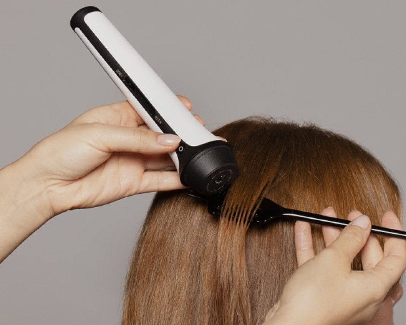 Osoba używa białego urządzenia do stylizacji włosów z czarnymi akcentami na brązowych włosach, przytrzymując pasmo czarnym grzebieniem.