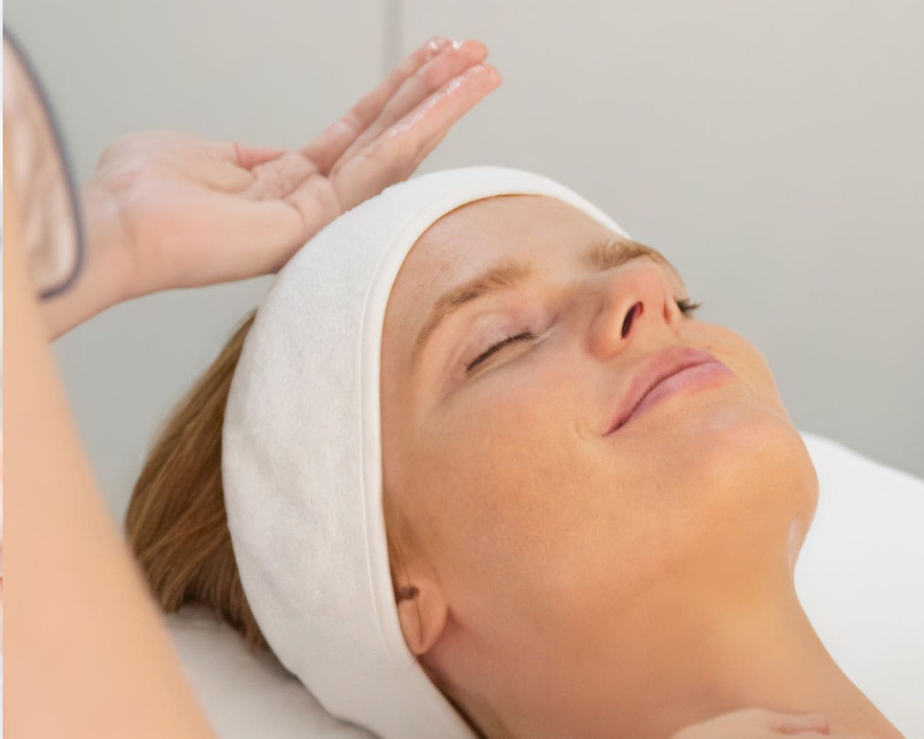 Kobieta z zamkniętymi oczami leży na białym ręczniku, na głowie ma białą opaskę, a ktoś nakłada jej coś na twarz.