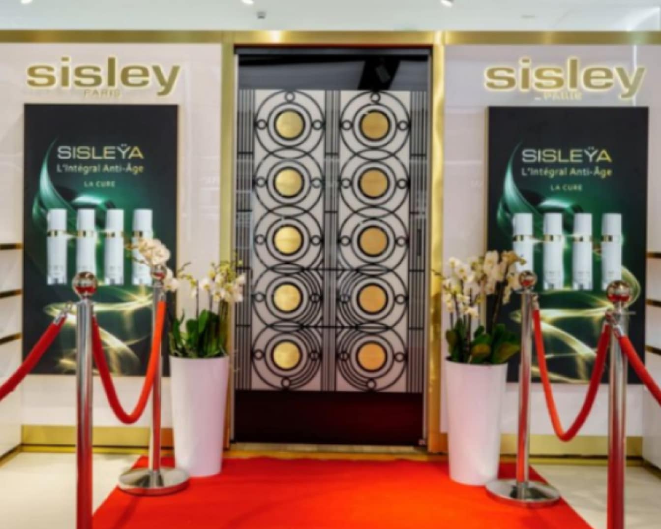 Wejście do stoiska Sisley Paris z czerwonym dywanem, złotymi akcentami i plakatami Sisleÿa L'Intégral Anti-Âge La Cure.