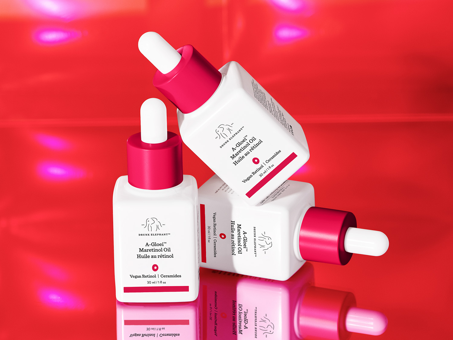 Nahaufnahme von drei weißen Drunk Elephant A-Gloei Maretinol Ölflaschen mit pinkfarbenen Deckeln und Pipetten auf einem glänzend roten Hintergrund, die Vegan Retinol und Ceramide enthalten.