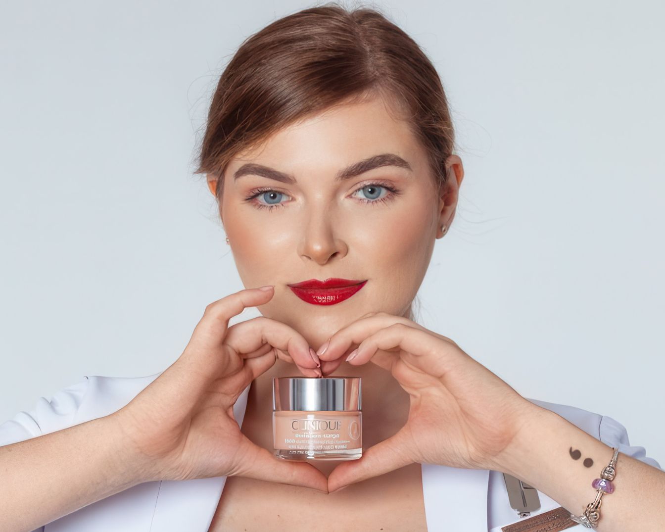 Kobieta z czerwoną szminką trzyma krem Clinique w przezroczystym słoiczku z srebrną nakrętką, układając dłonie w kształt serca.