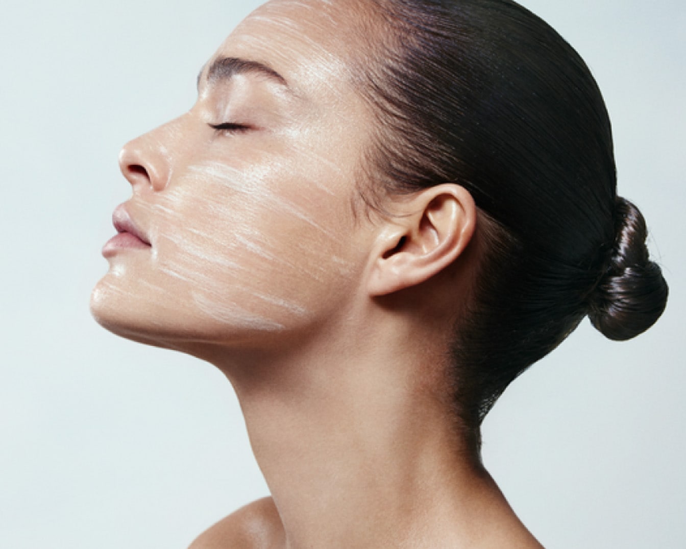 Profil kobiety z zamkniętymi oczami i włosami upiętymi w kok, na twarzy ma nałożoną białą maseczkę.