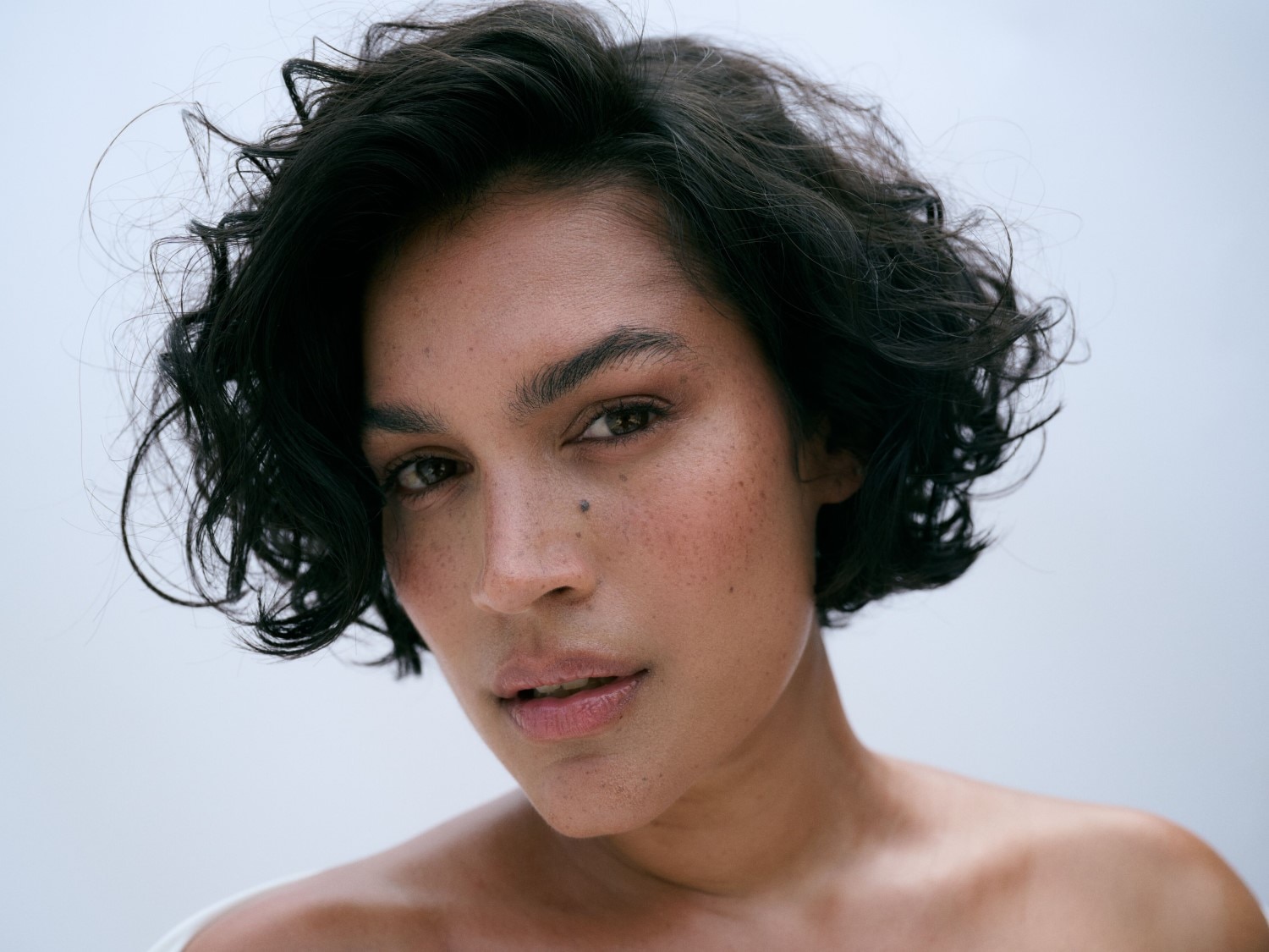 Nahaufnahme einer Frau mit dunklen, gewellten Haaren, Sommersprossen und einem Muttermal im Gesicht, die direkt in die Kamera schaut.