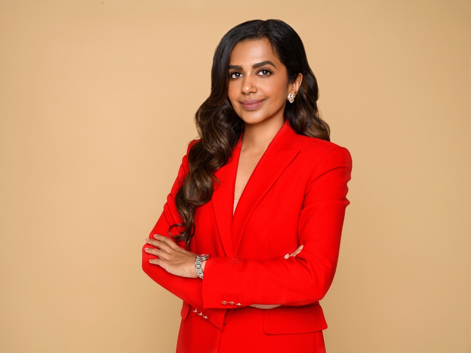 Eine Frau mit dunklen, gewellten Haaren und einem roten Blazer posiert selbstbewusst vor einem beigen Hintergrund.