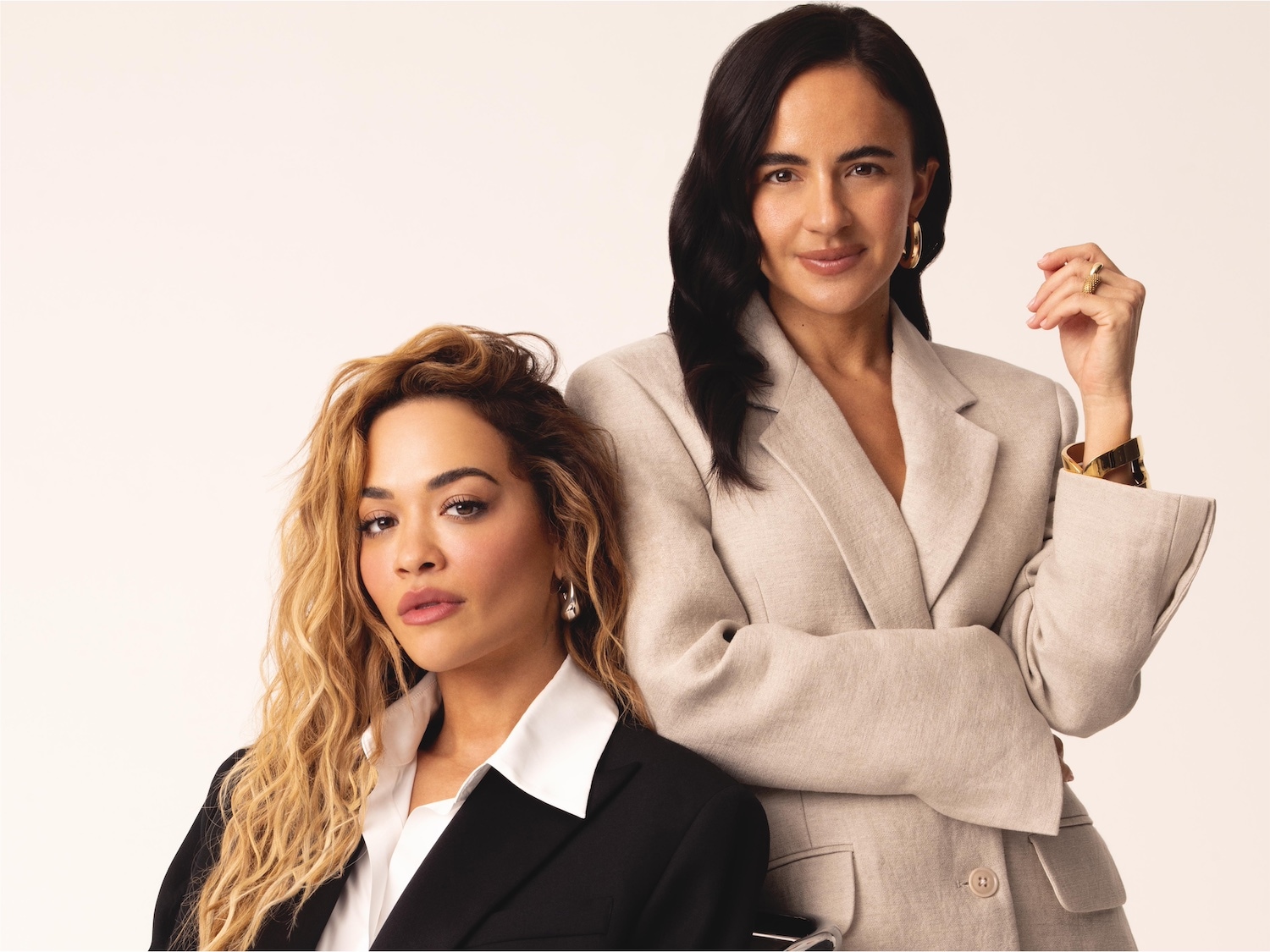 Zwei Frauen posieren selbstbewusst: Eine mit blonden Locken im schwarzen Blazer, die andere mit dunklen Haaren im beigen Blazer, beide strahlen Stärke und Eleganz aus.