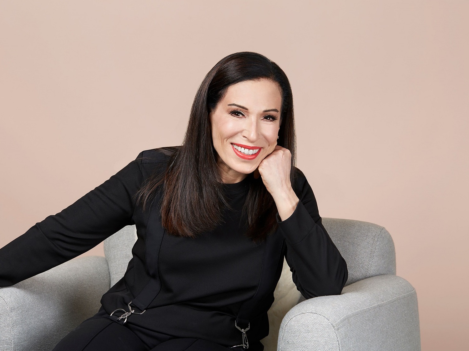 Eine lächelnde Frau mit dunklen, glatten Haaren und rotem Lippenstift sitzt in einem grauen Sessel, ihr Gesicht ruht auf ihrer Hand.