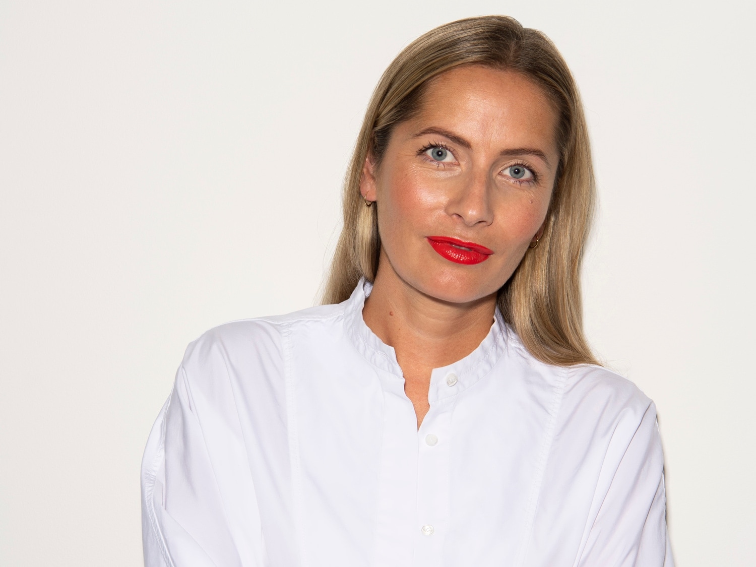 Porträt einer Frau mit blonden, glatten Haaren, blauen Augen und roten Lippen, die ein weißes Hemd trägt.