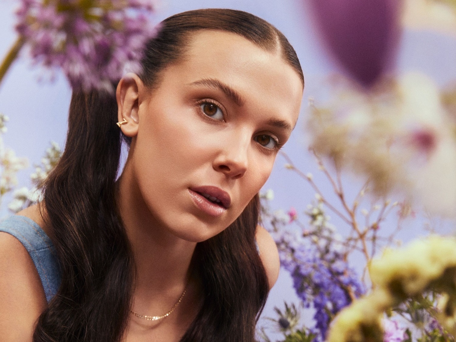 Nahaufnahme einer jungen Frau mit braunen Haaren, die zu Zöpfen gebunden sind, mit einem goldenen Ohrring und einer goldenen Halskette, umgeben von Blumen.