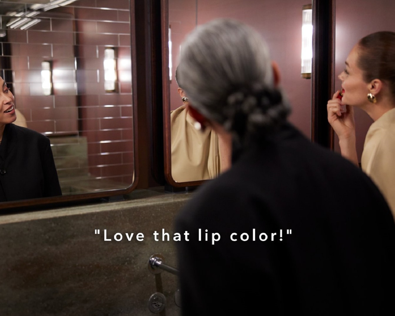 Kobieta w beżowej bluzce maluje usta czerwoną szminką przed lustrem, a druga kobieta w czarnej marynarce komplementuje kolor szminki.