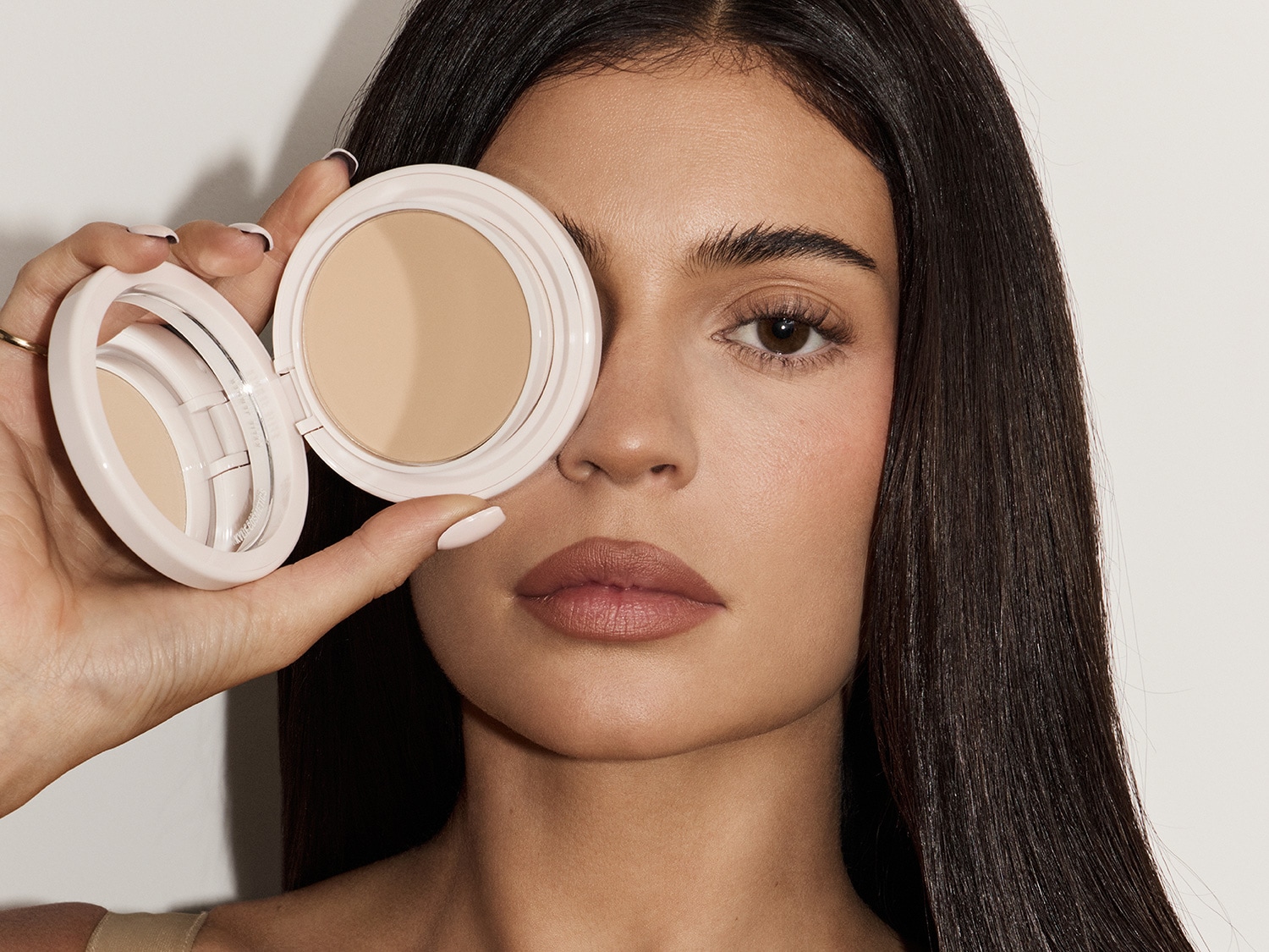 Eine Frau mit langen, dunklen Haaren hält eine geöffnete, runde, hellrosa Puderdose vor ihr Auge, die einen Spiegel und hellbeige Puder enthält.