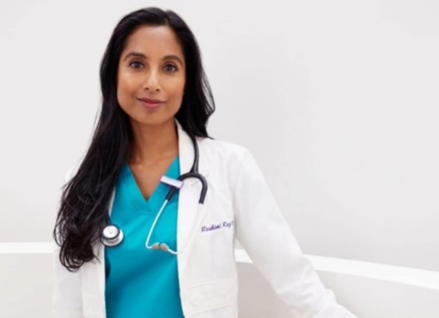 Eine Ärztin mit dunklen, langen Haaren trägt einen weißen Kittel über einem türkisfarbenen Oberteil und ein Stethoskop um den Hals.