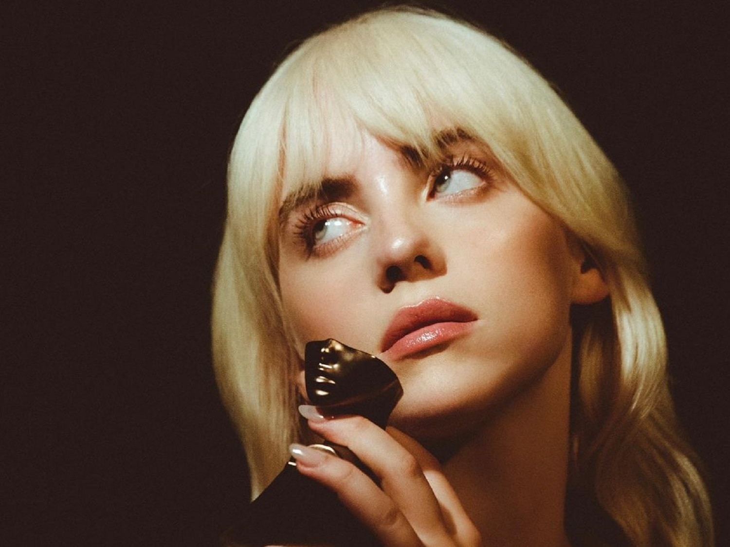 Nahaufnahme von Billie Eilish mit blonden Haaren, die einen schwarzen Parfümflakon mit goldfarbenem Verschluss hält und verträumt nach oben blickt.