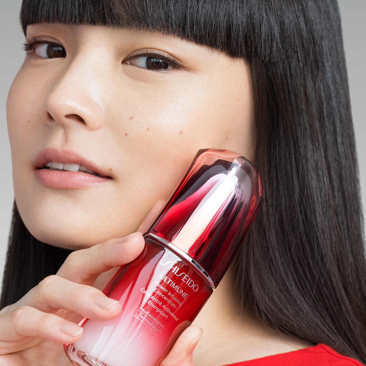 Een vrouw met donker haar houdt een rode Shiseido Ultimune Power Infusing Concentrate fles tegen haar gezicht.