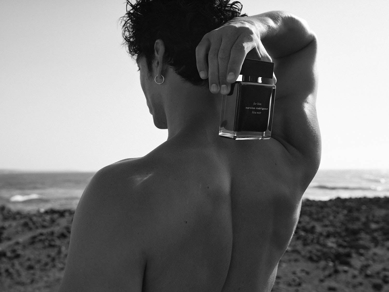 Ein Mann mit freiem Oberkörper hält eine Flasche Narciso Rodriguez Bleu Noir Duft vor dem Hintergrund eines sonnigen Strandes.