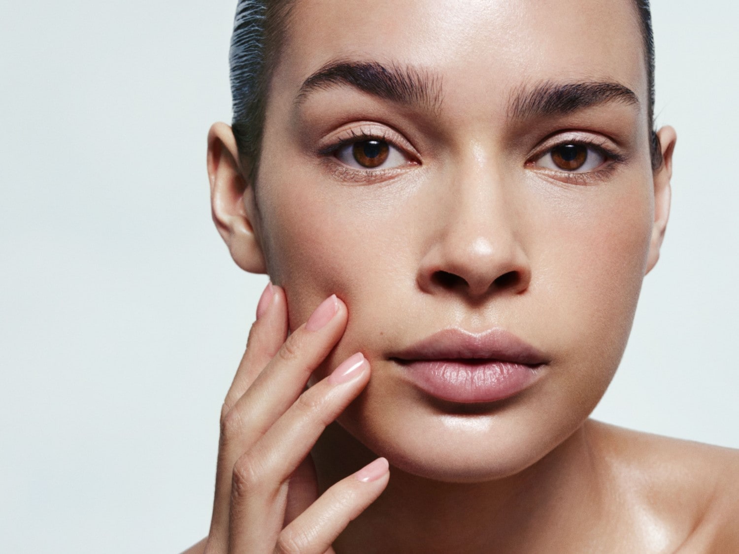 Gros plan sur le visage d'une femme à la peau éclatante, les cheveux tirés en arrière, touchant délicatement sa joue avec des ongles manucurés rose pâle.
