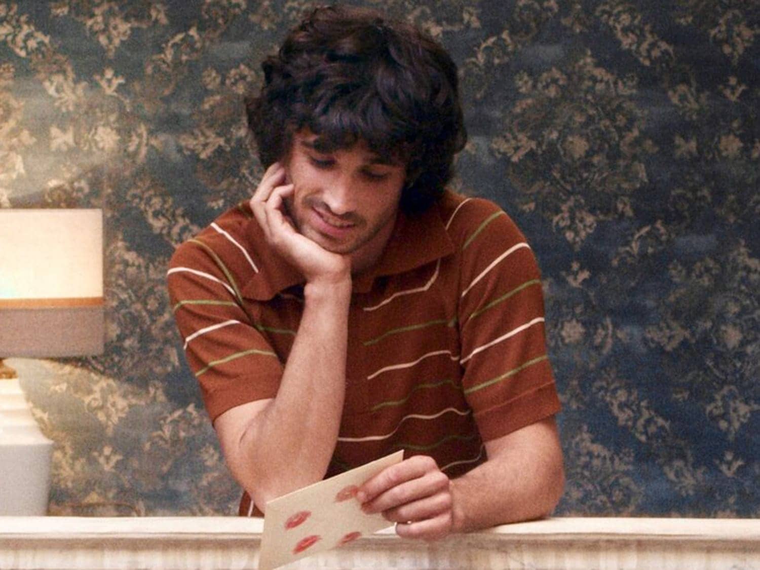 Mladý muž s tmavými kudrnatými vlasy se opírá o ruku a s úsměvem si prohlíží dopis s otisky rtů.