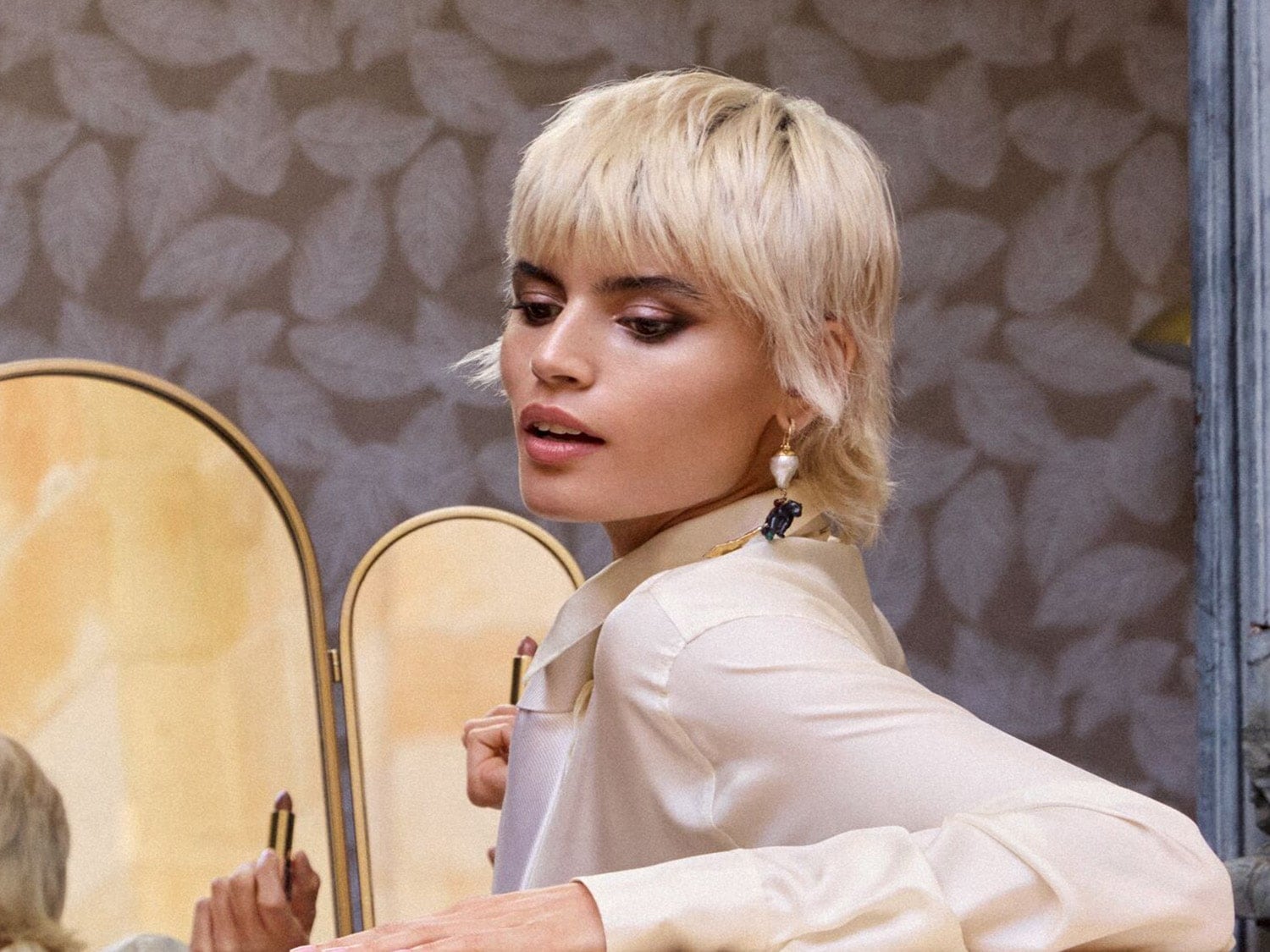 Blondýna s krátkými vlasy nanáší rtěnku před zrcadlem, má na sobě saténovou košili a výrazné náušnice.