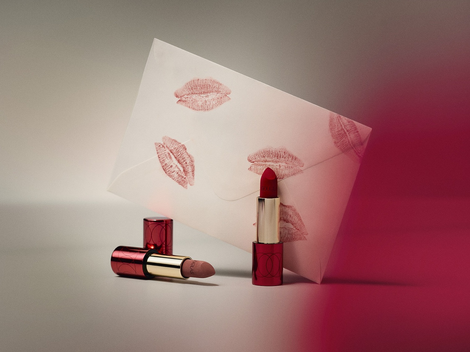 Červený rúž so zlatým obalom stojí vedľa bielej obálky s odtlačkami pier, pripomínajúci tajomstvo krásy a elegancie.