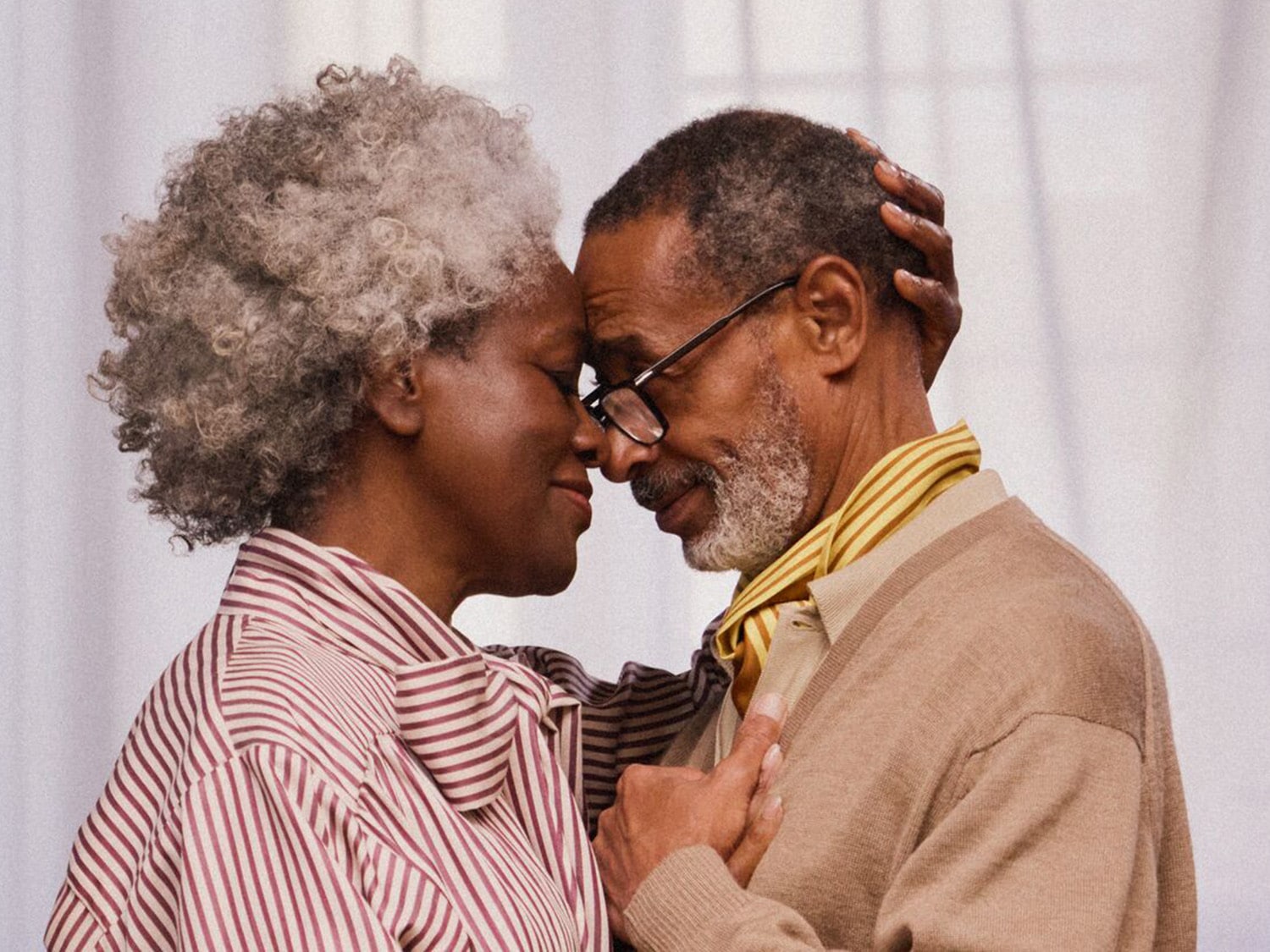 Starší pár sa nežne dotýka čelami, žena s kučeravými sivými vlasmi v pruhovanej blúzke a muž s okuliarmi v béžovom svetri, držia sa za ruky.