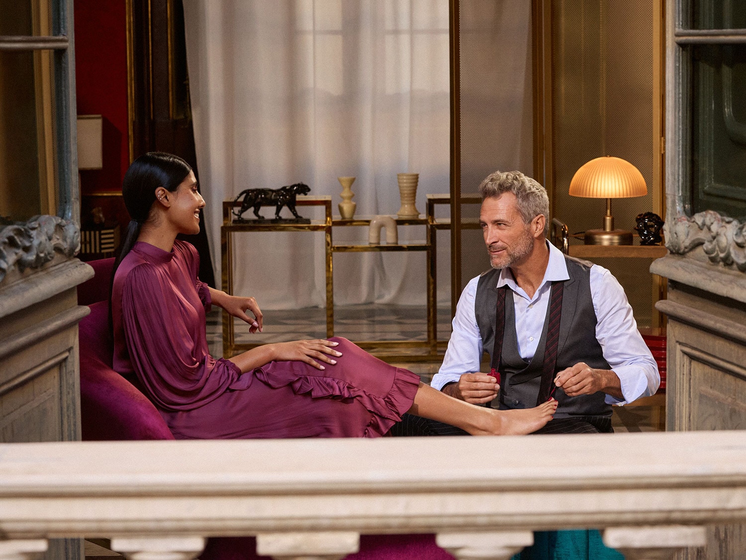 Muž v sivom obleku s červenou fľaštičkou laku na nechty lakuje žene nechty na nohách, ktorá sedí oproti nemu v bordových šatách.