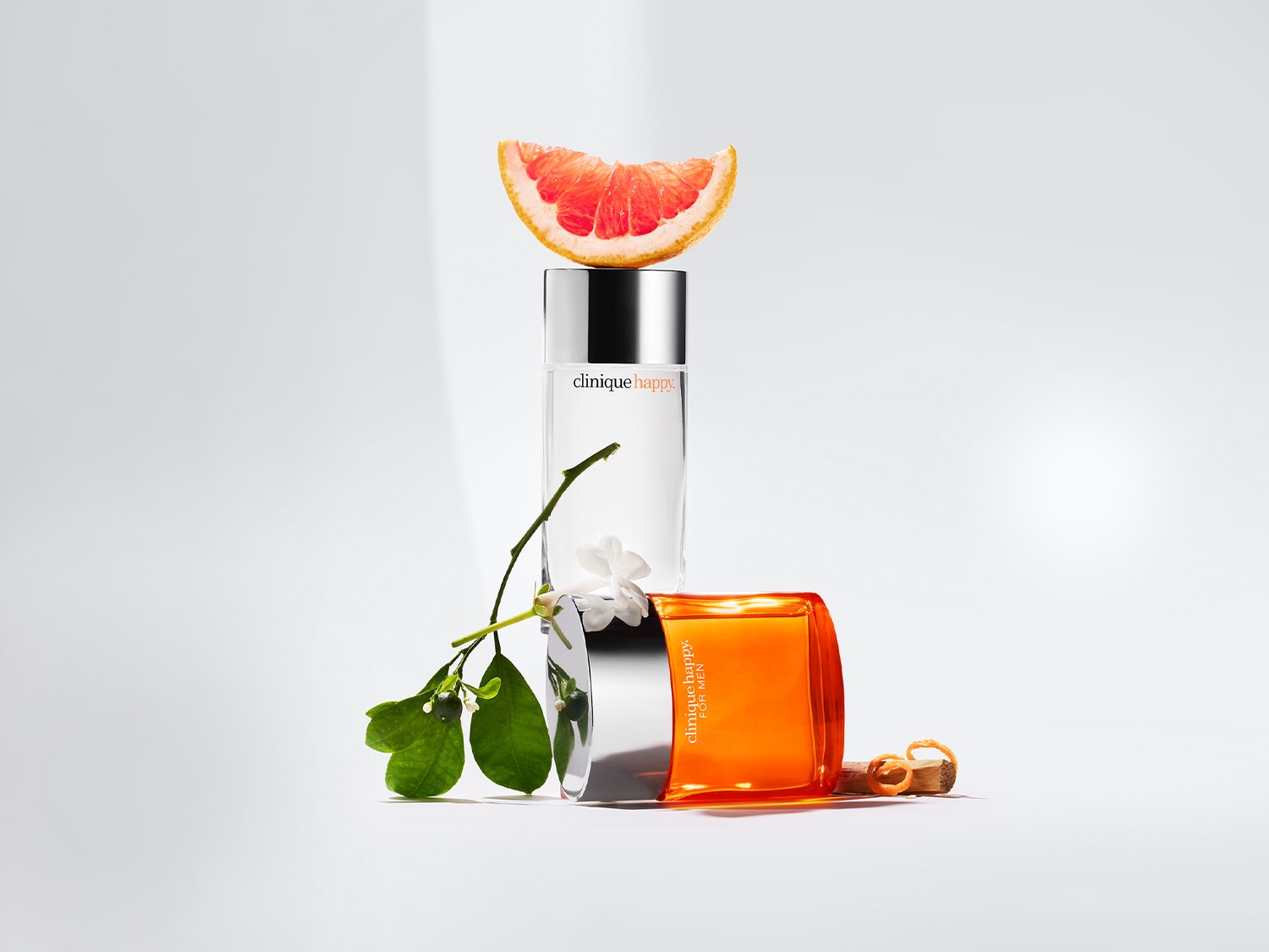 Clinique Happy und Clinique Happy for Men Duftkollektion, bestehend aus einem transparenten Flakon mit silbernem Verschluss, einem orangefarbenen Flakon, einer Grapefruitscheibe und einem Blumenarrangement.
