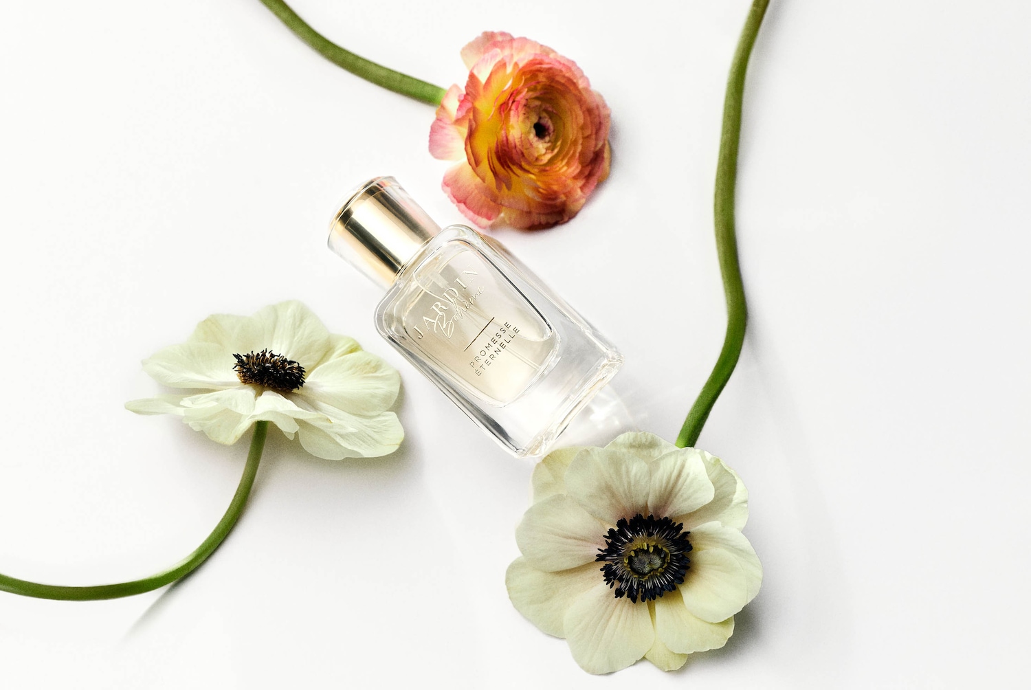 Skaidraus stiklo kvepalų buteliukas su auksiniu dangteliu, apsuptas baltų ir oranžinių žiedų ant balto paviršiaus.