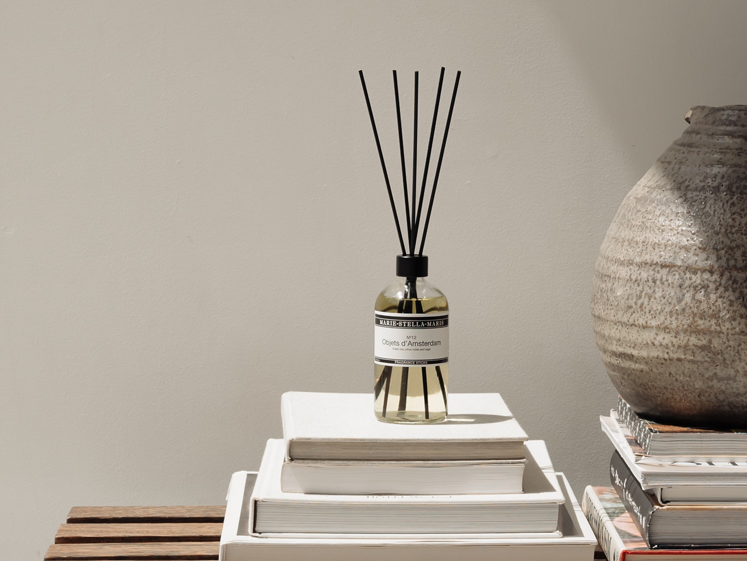 Een Marie-Stella-Maris geurstokjes diffuser staat op een stapel witte boeken, met zwarte stokjes die uit de fles steken, naast een rustieke stenen vaas.