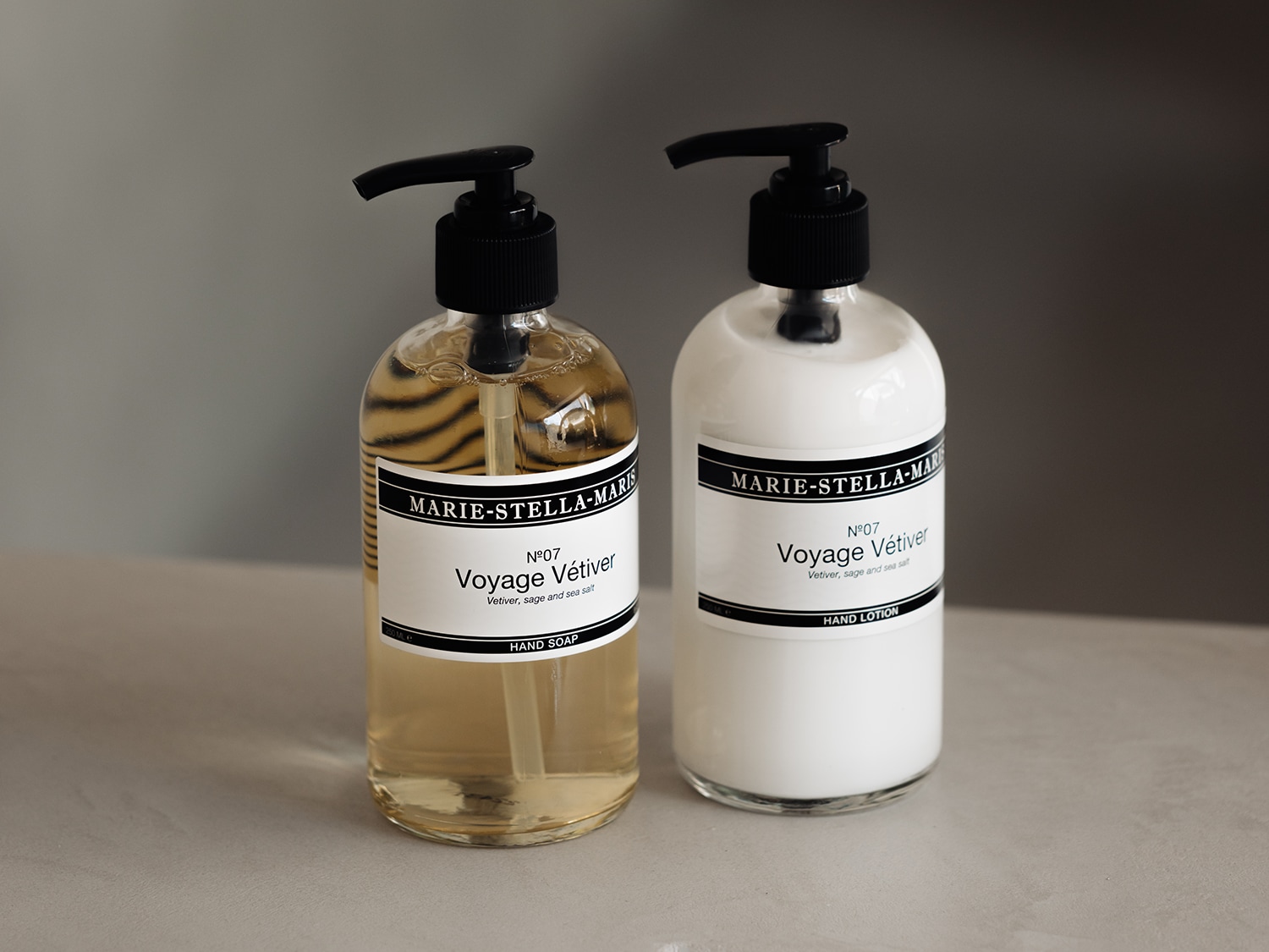Twee glazen flessen met zwarte pompen, de ene gevuld met handzeep en de andere met handlotion van Marie-Stella-Maris N°07 Voyage Vétiver, met de geur van vetiver, salie en zeezout.