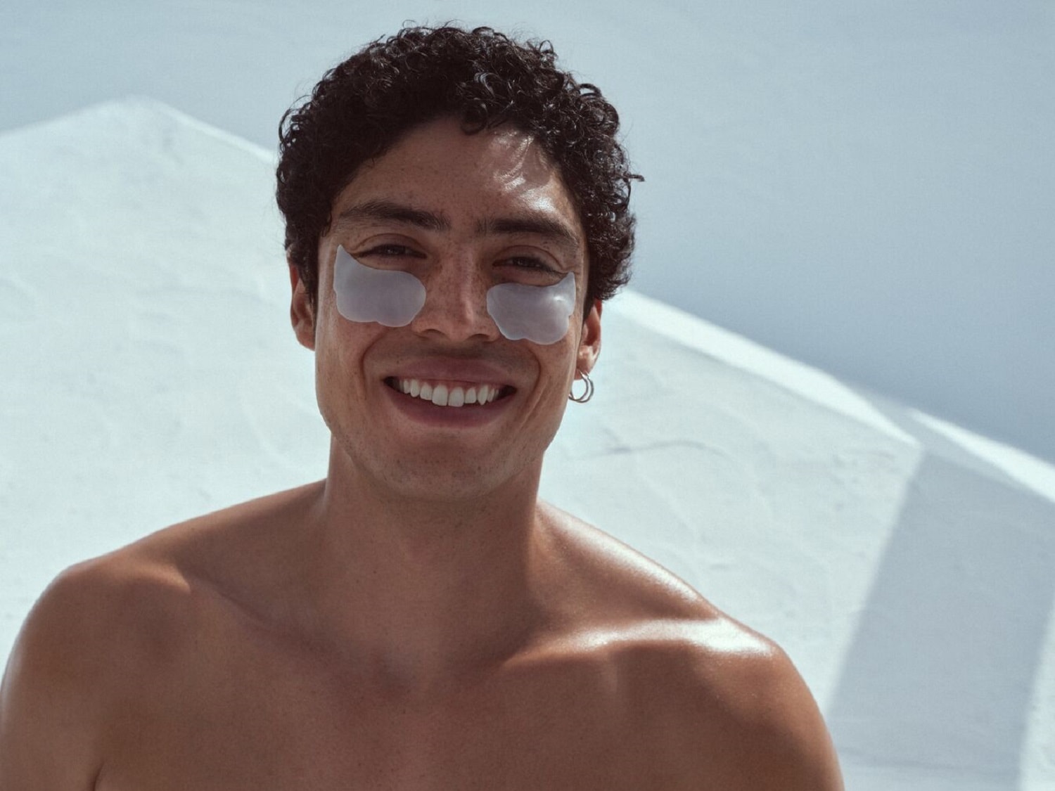 Šypsodamasis jaunas vyras su tamsiais garbanotais plaukais ir sidabriniais auskarais ant skruostų užsidėjęs skaidrius paakių pleistrus.