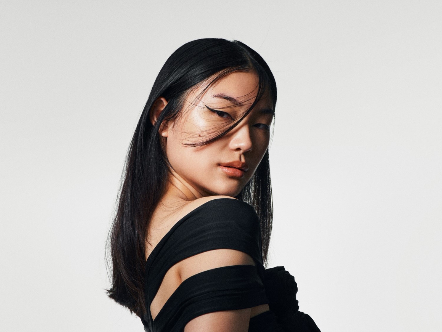 Een vrouw met lang, zwart haar en een zwarte jurk kijkt over haar schouder met een subtiele eyeliner.