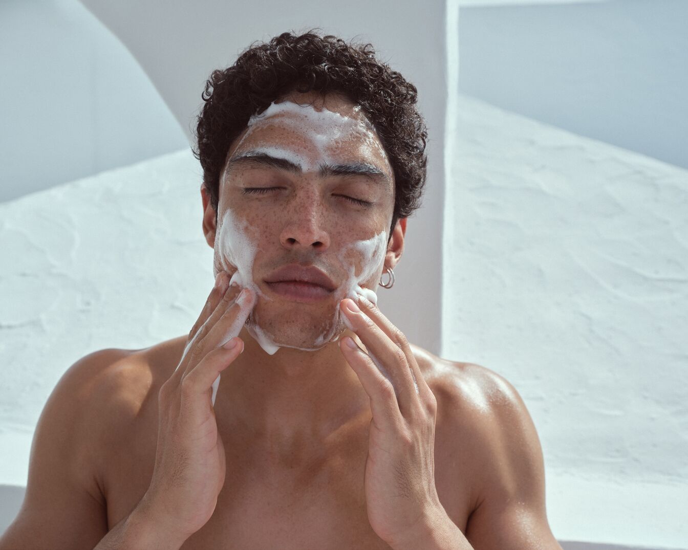 Ein Mann mit lockigem Haar und geschlossenen Augen massiert sein Gesicht mit einer weißen, schaumigen Reinigungslotion.
