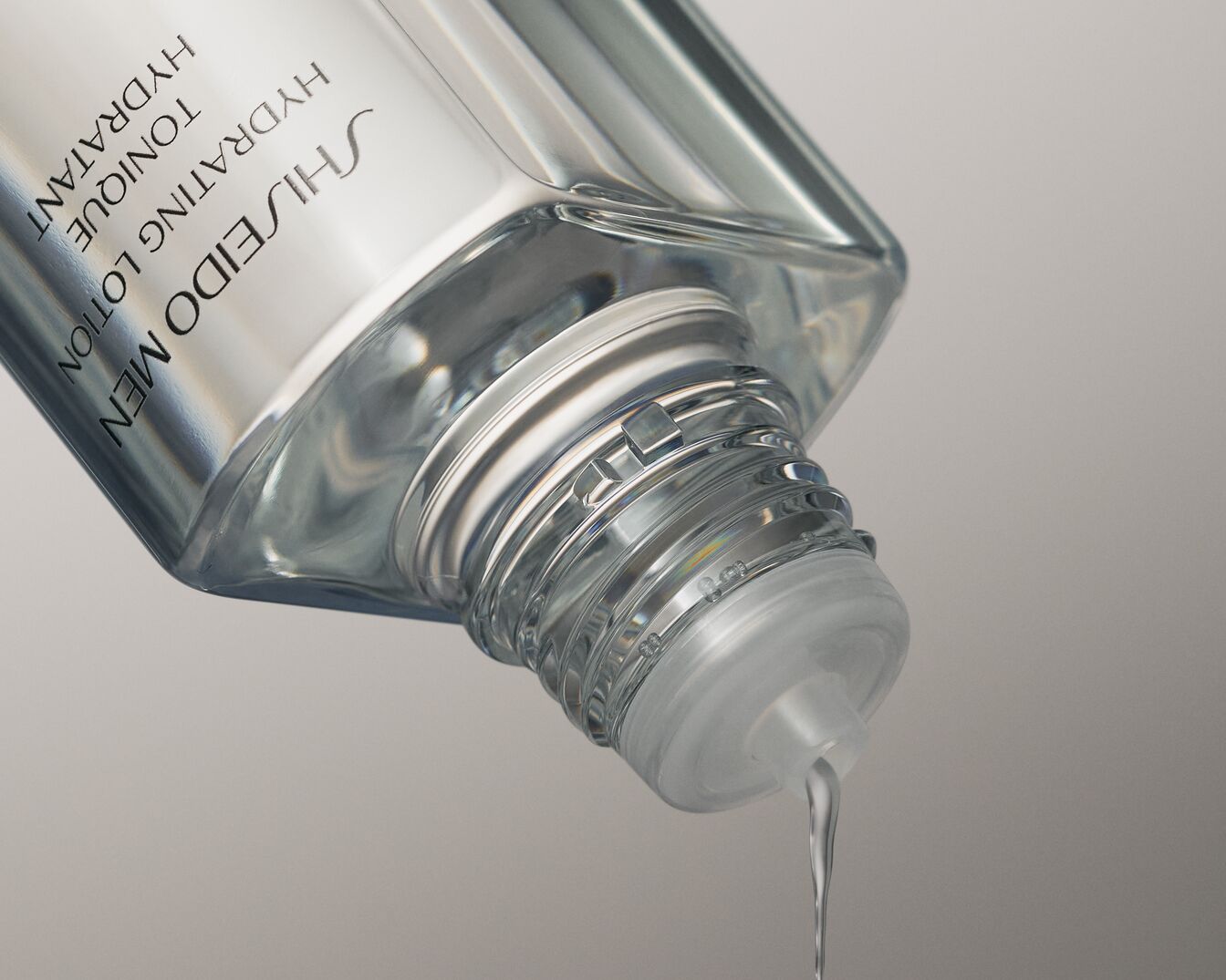 Nahaufnahme einer durchsichtigen Flasche Shiseido Men Hydrating Lotion, aus der ein klarer Tropfen auf hellgrauem Hintergrund fließt.