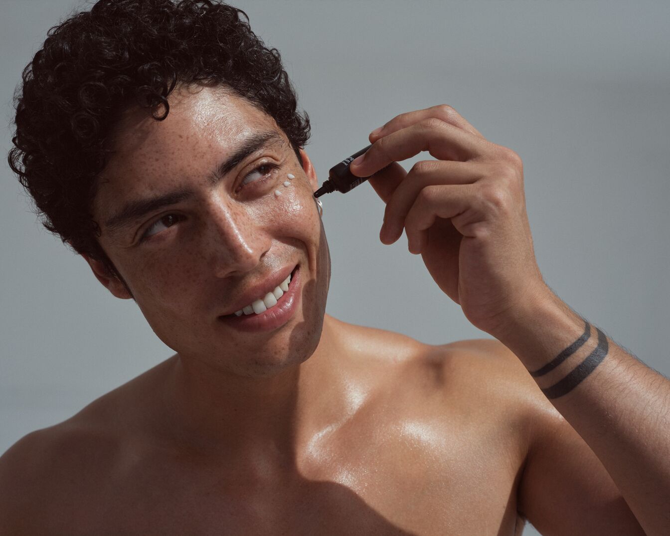 Ein Mann mit lockigem Haar und sonnengebräunter Haut trägt eine kleine Menge weißer Creme aus einer dunkelbraunen Tube auf seine Wange auf.