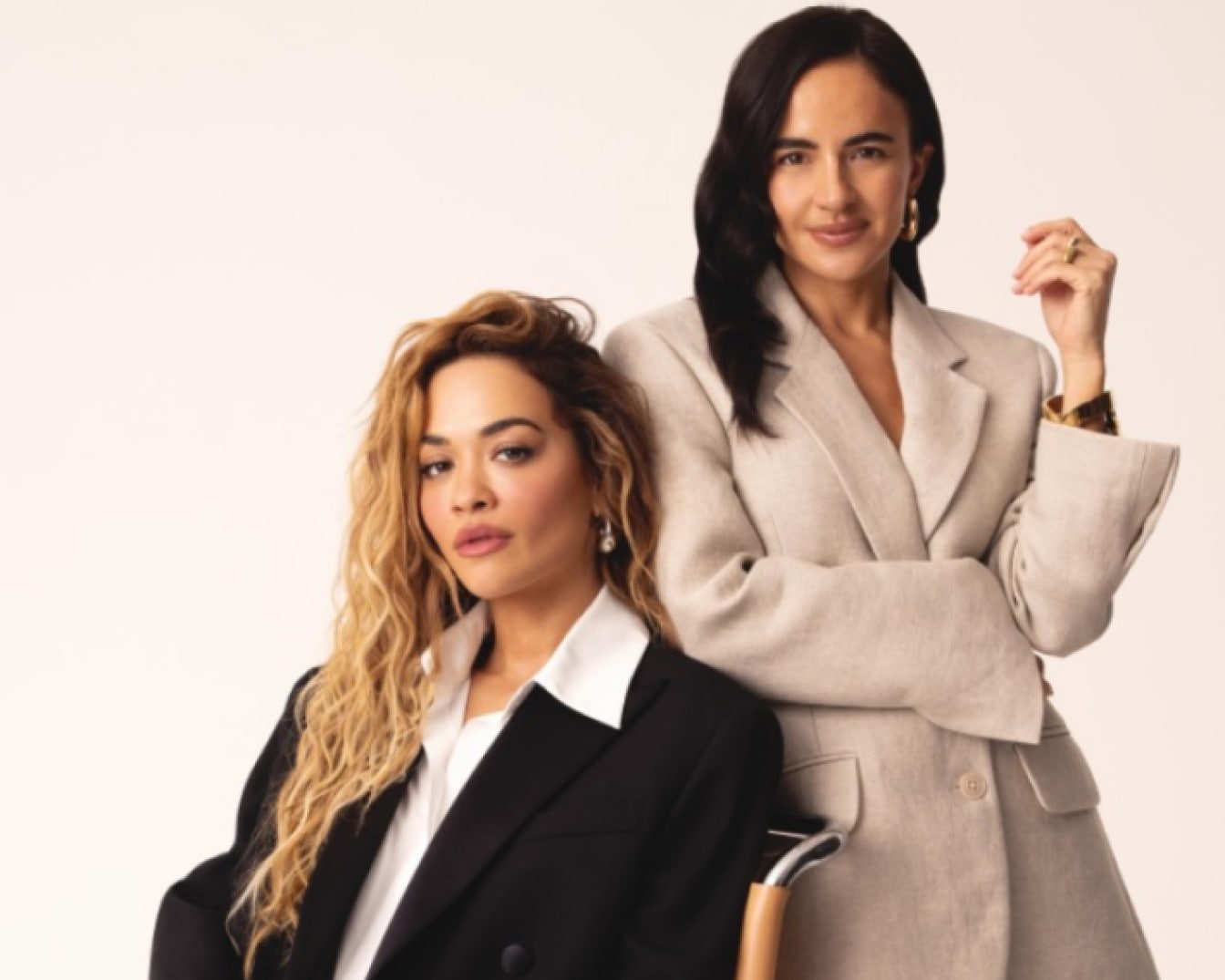 Šviesiaplaukė moteris juodu švarku sėdi šalia tamsiaplaukės moters su smėlio spalvos švarku, abi atrodo pasitikinčios ir stilingos.